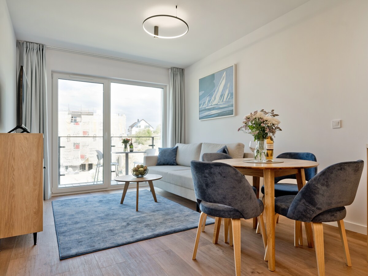 A modern living area features a light grey sofa and a round wooden dining table surrounded by four upholstered chairs. A textured rug complements the space. Large glass doors provide access to a balcony, allowing natural light to fill the room.
