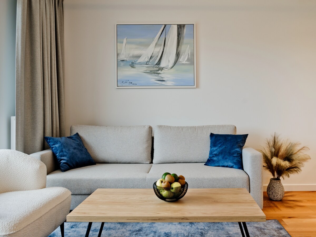 A light-filled living area features a soft gray sofa adorned with blue accent pillows. A wooden coffee table sits in the center, hosting a bowl of fresh fruit. A framed painting of sailboats is displayed on the wall, accompanied by a decorative plant in the corner.