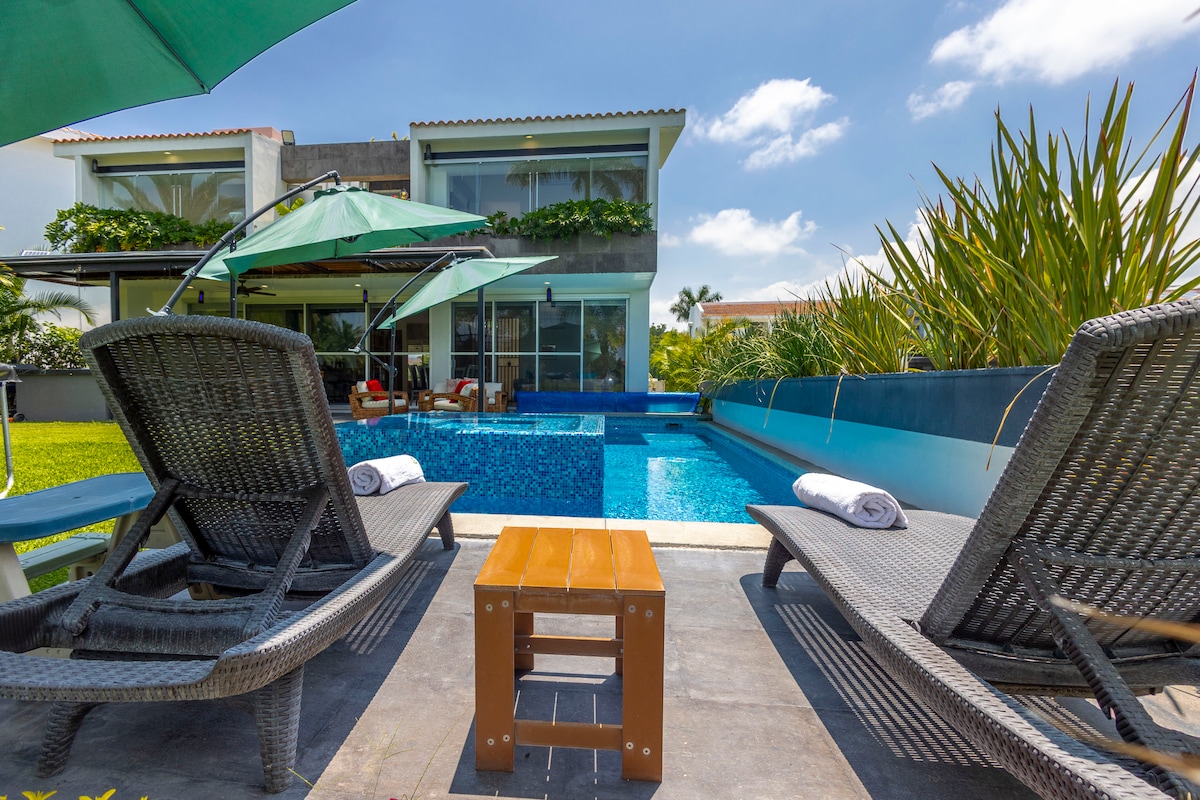 Two lounge chairs are positioned beside a sparkling blue pool, surrounded by vibrant greenery. A small table stands between the chairs. Large windows reflect the exterior of a modern two-story house, with umbrellas providing shade overhead.