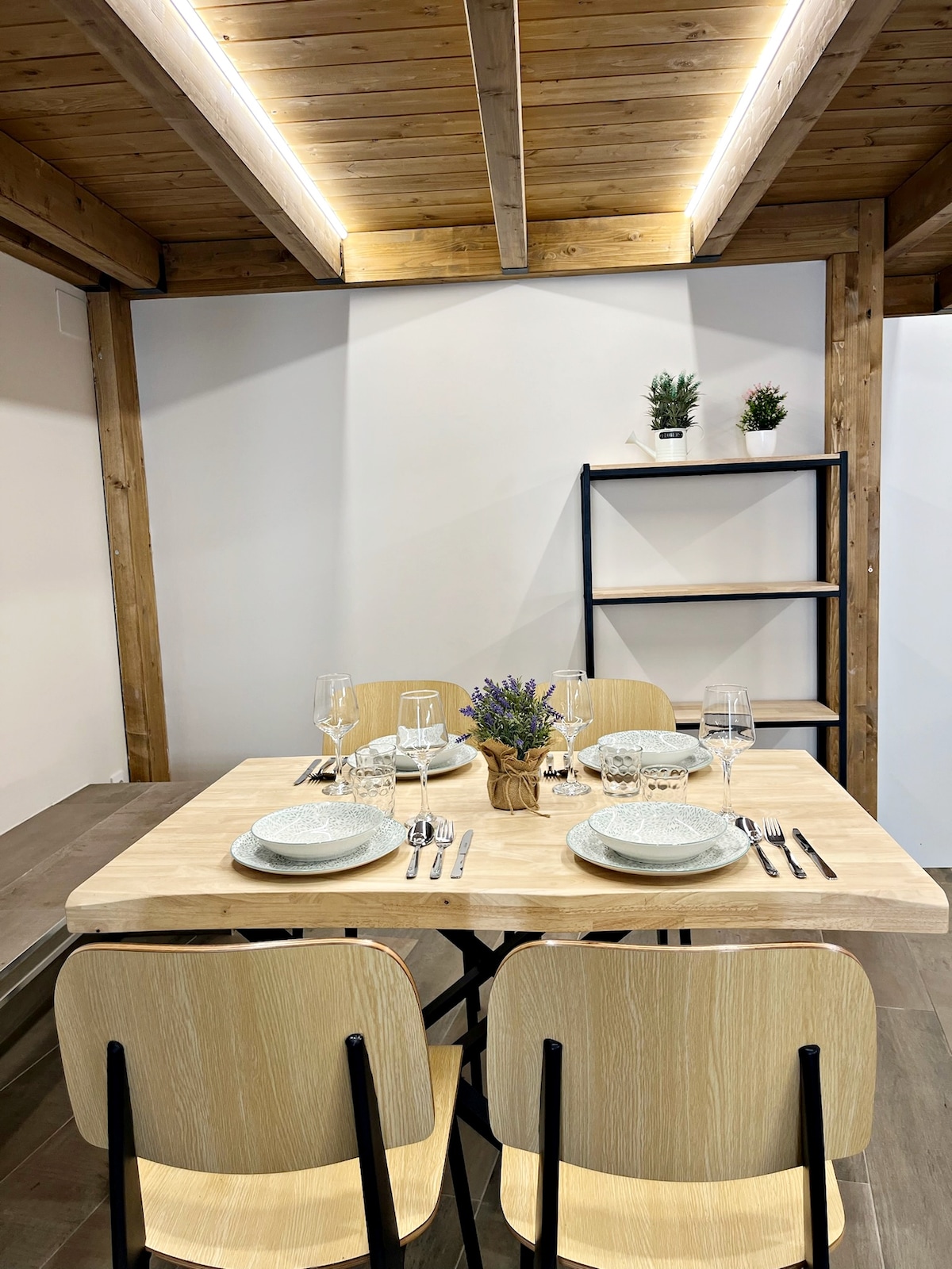 A dining area features a wooden table set for four, complete with plates, glasses, and silverware. Light wood chairs surround the table. A decorative plant and a small vase of lavender add a touch of color against the backdrop of bright, minimalist walls and wooden beams.