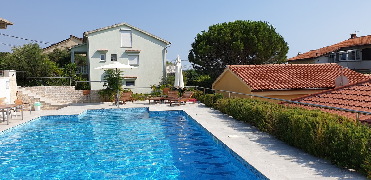 A shared outdoor pool is surrounded by sun loungers and greenery, providing a relaxing space. A two-story house stands nearby, featuring light-colored walls and white window frames. The scene is framed by a clear blue sky and the rooftops of adjacent buildings.