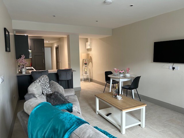 A modern living area features a comfortable sofa draped with a teal throw, while a coffee table sits in front. A dining table for two is positioned nearby, and a wall-mounted television adds convenience. The space is well-lit with neutral-colored walls.