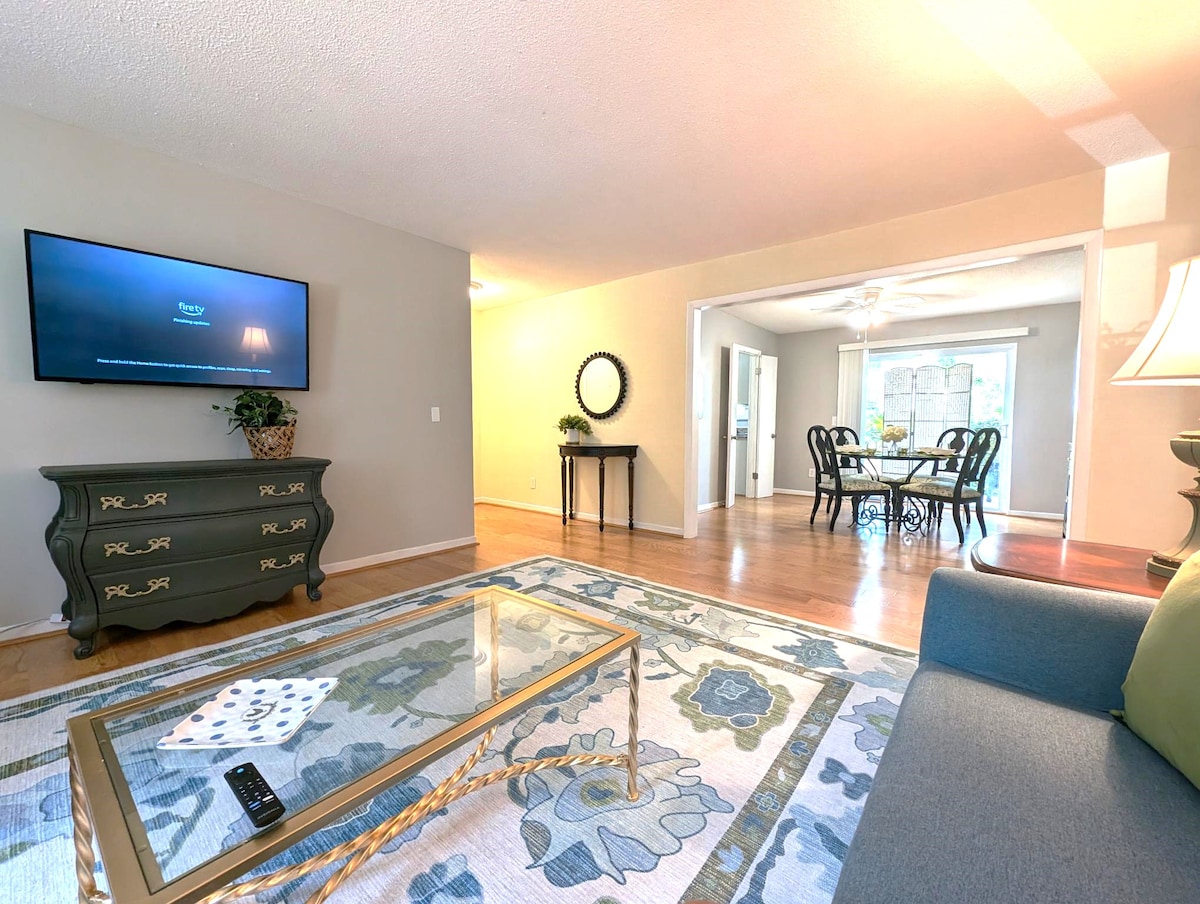 A welcoming living area is displayed, featuring a comfortable sofa beside a decorative coffee table. A flat-screen television is mounted on the wall, and natural light enters through a nearby window, illuminating the room. A dining space is visible in the background.