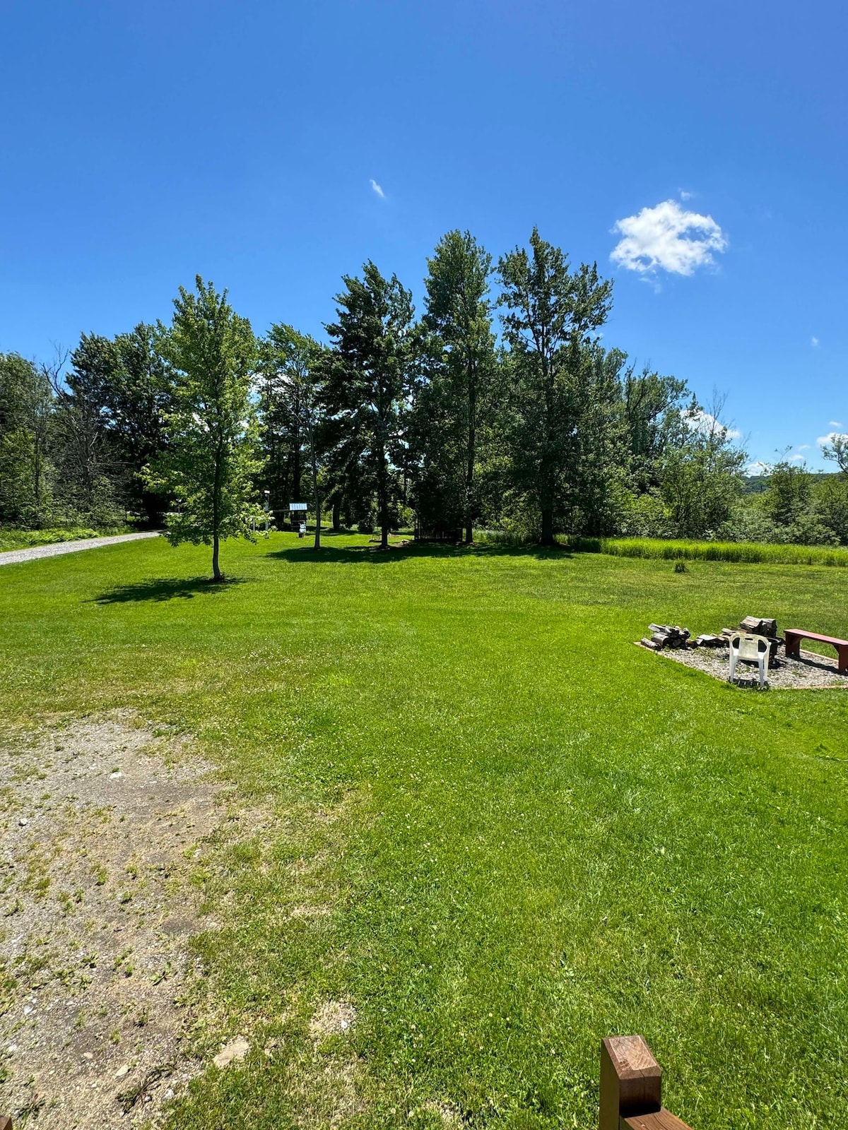 A spacious outdoor area is surrounded by greenery, featuring a well-maintained lawn and several trees. A fire pit is located on the ground, with a walking path visible leading away into the trees. The sky is clear and blue, contributing to a serene atmosphere.