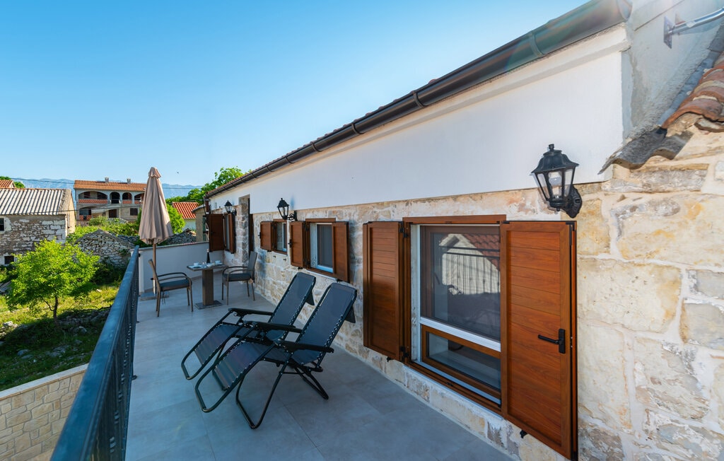 A spacious terrace is adorned with two lounge chairs and a small table, offering a comfortable spot to relax. Wooden shutters frame the large window, while sunlit stone walls add rustic character. Lush greenery can be seen in the background, enhancing the serene outdoor setting.