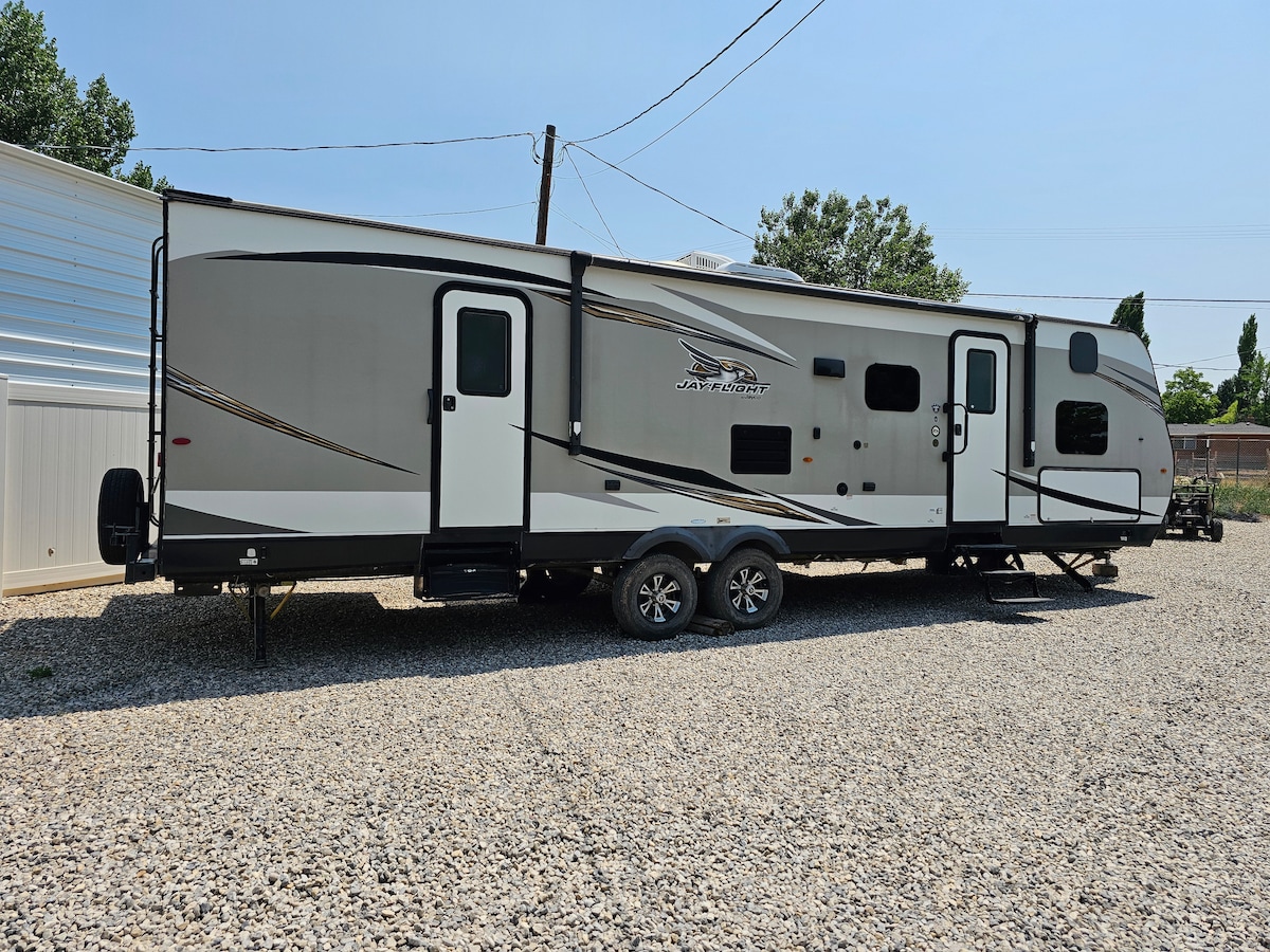 A spacious travel trailer is parked on gravel, featuring an exterior in neutral tones with sleek lines and subtle detailing. The trailer is equipped with multiple windows and a streamlined design, surrounded by a natural setting with open space in the background.