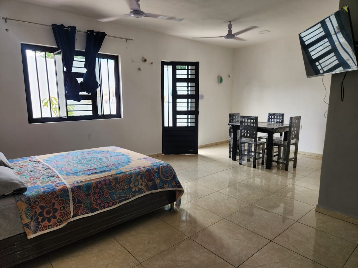 The room features a spacious bed covered with a patterned throw. A dining table with four chairs is positioned near a window, and a wall-mounted television adds convenience. Light-colored walls and tiled flooring create a bright and open atmosphere.