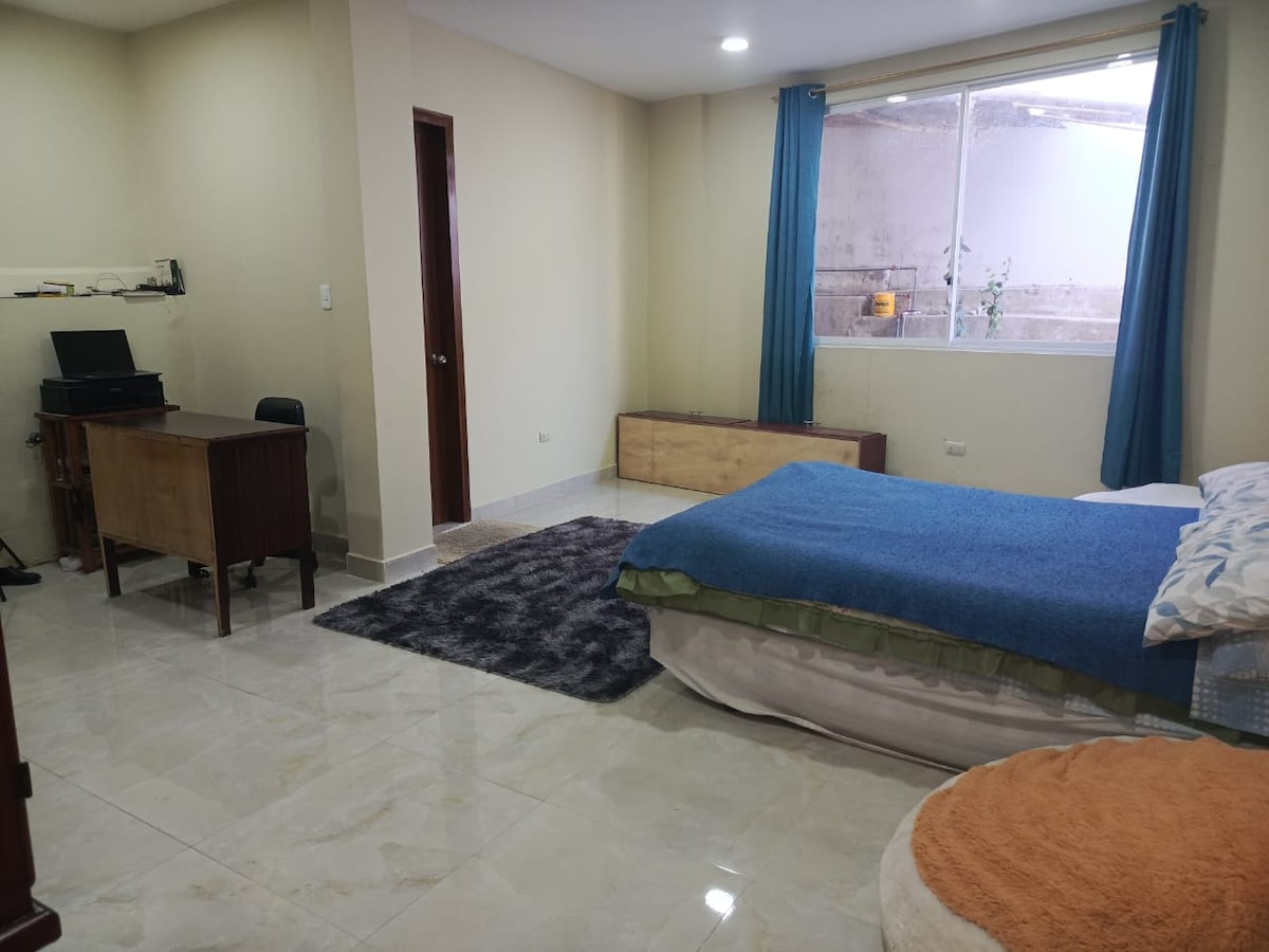 A spacious bedroom features a large bed covered with a blue blanket and white sheets. A wooden desk and chair are positioned against one wall. Natural light enters through a large window, complemented by light blue curtains. A soft rug is placed on the floor.