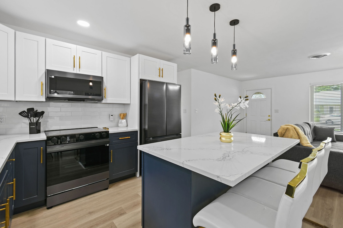 A modern kitchen is presented with sleek cabinetry in white and navy tones. A spacious island with a marble countertop offers seating for four, complemented by stylish gold accents. Stainless steel appliances are featured, including an oven and refrigerator, enhancing the practical layout for family gatherings.