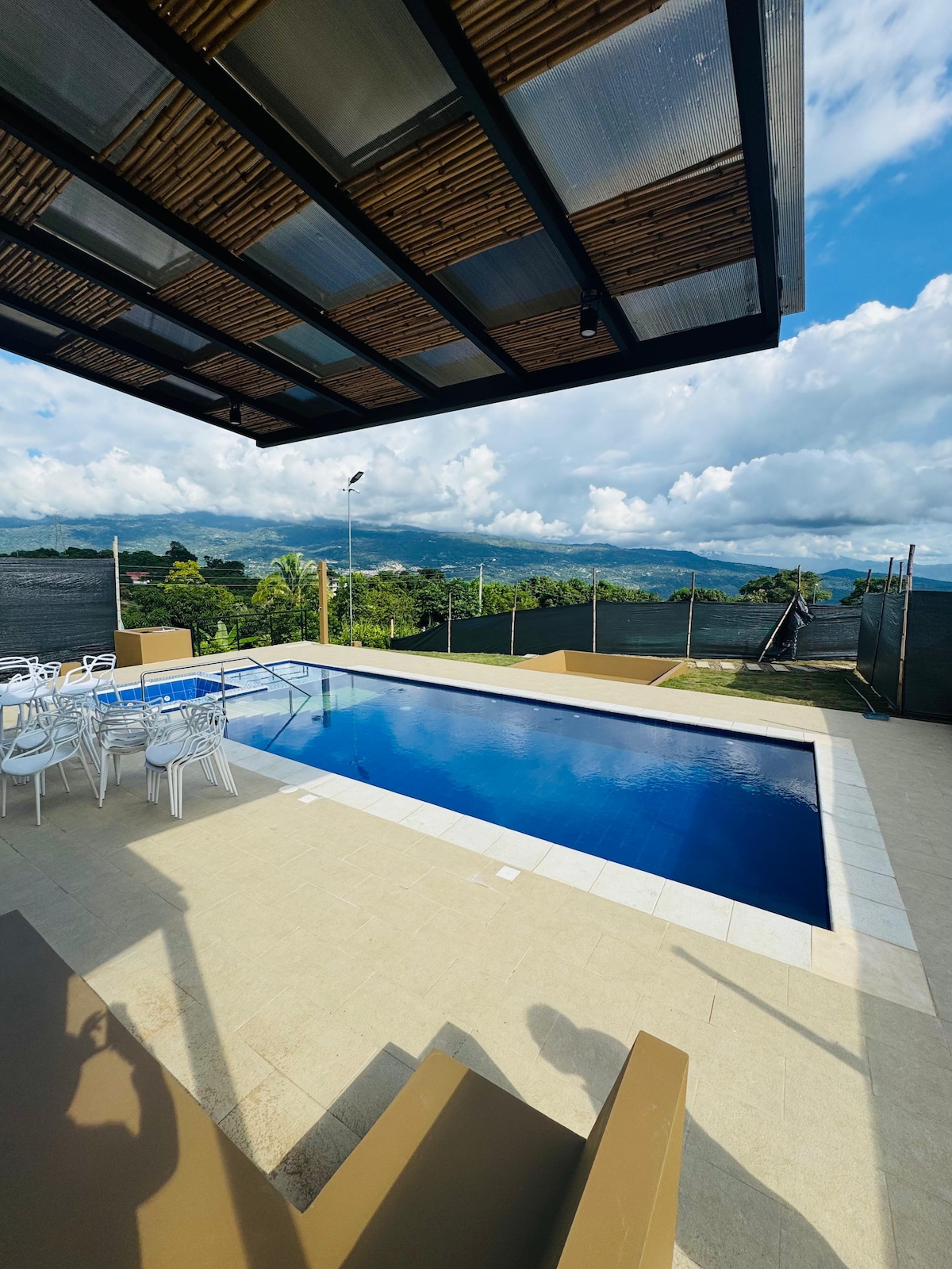 A contemporary outdoor space features a swimming pool with a clear blue surface, surrounded by a light-colored deck. An area with white chairs is positioned nearby, complemented by a shaded pergola overhead. Lush greenery and distant mountains create a serene backdrop.