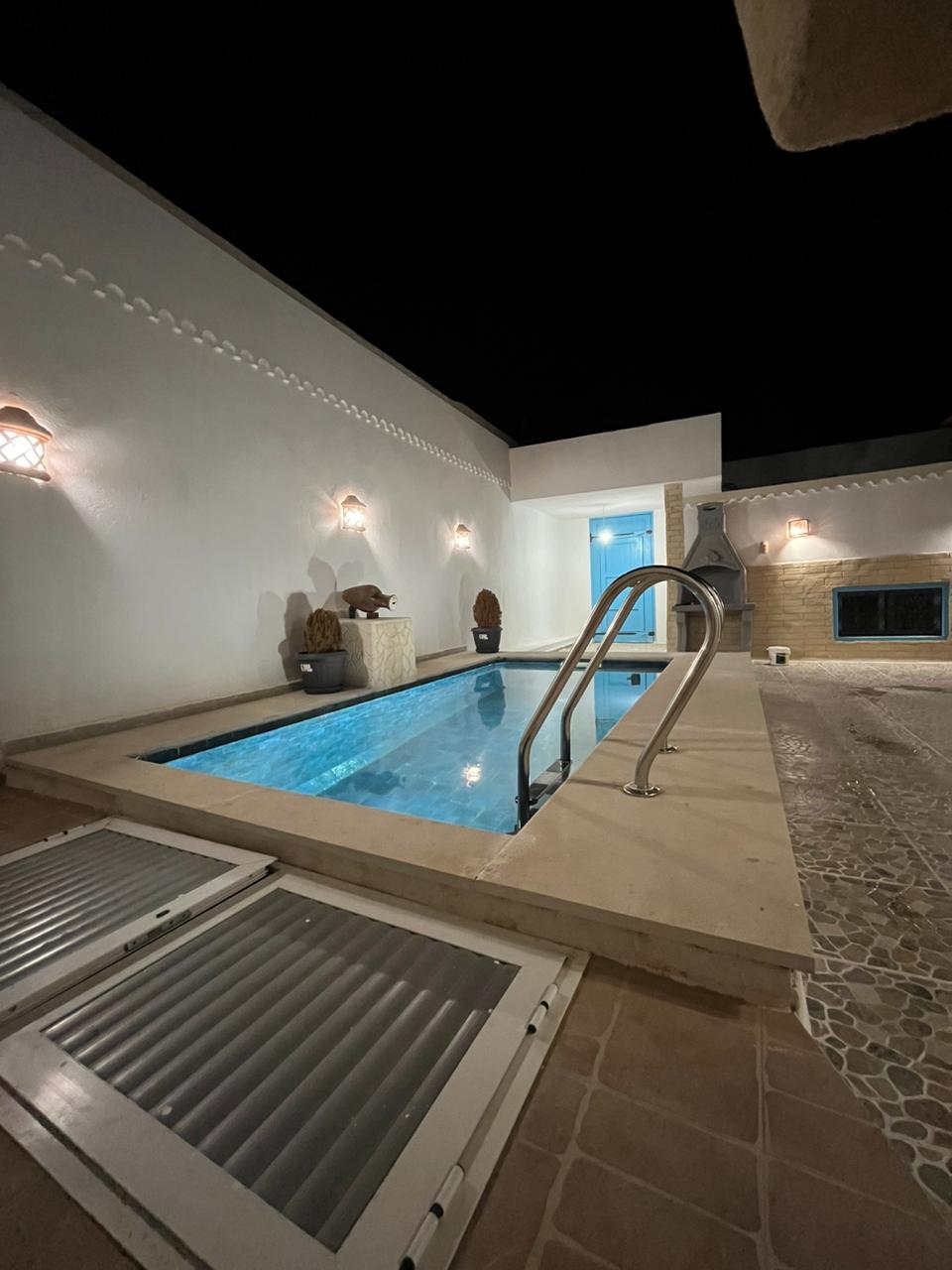 A private swimming pool is surrounded by a well-lit terrace, featuring elegant wall sconces that enhance the serene atmosphere. The pool is showcased with a set of stainless steel steps leading into the water, while a fireplace is visible in the background.