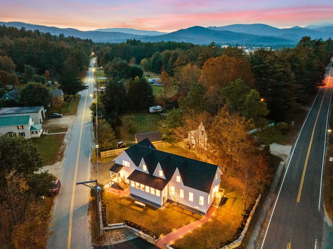 An aerial view captures the home illuminated by warm exterior lights, set against a backdrop of mountains and a colorful sunset. The road lines the property, with trees and neighboring homes visible, creating a serene and inviting atmosphere in the heart of nature.
