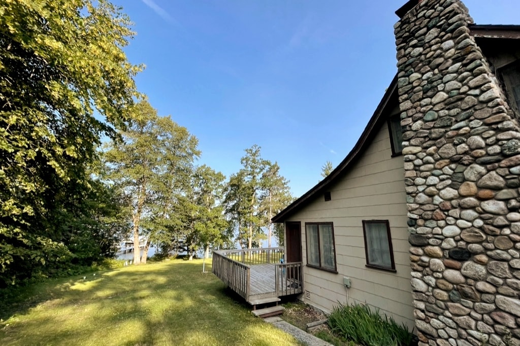 A side view of the cottage is presented, showcasing a rustic stone chimney and a wooden deck. Lush green grass surrounds the cottage, with trees providing natural shade and the lake visible in the background, suggesting a serene outdoor environment.