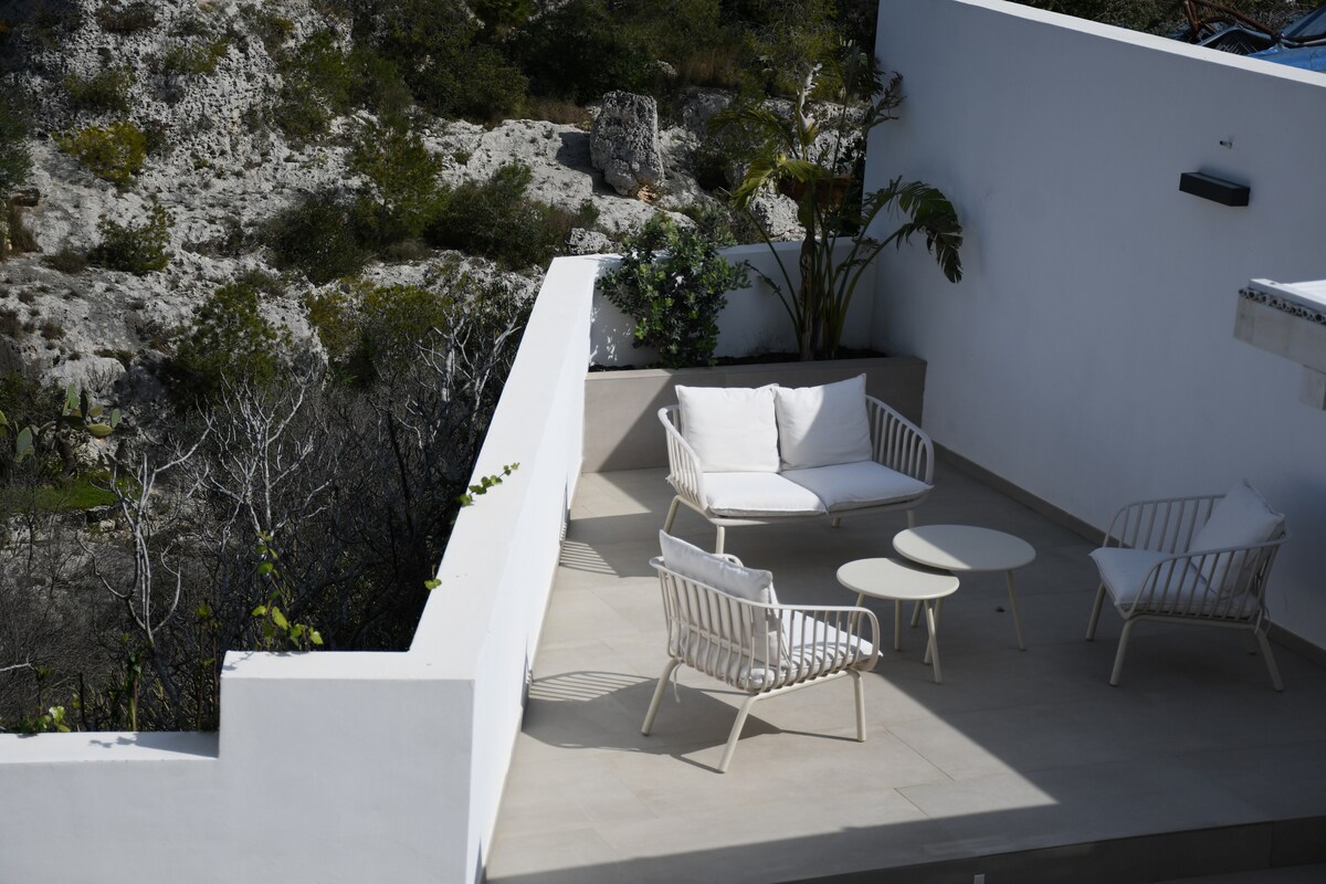 An outdoor area features a modern seating arrangement consisting of a white sofa and two armchairs surrounding a circular table. The space is framed by greenery, and a rocky landscape is visible nearby.