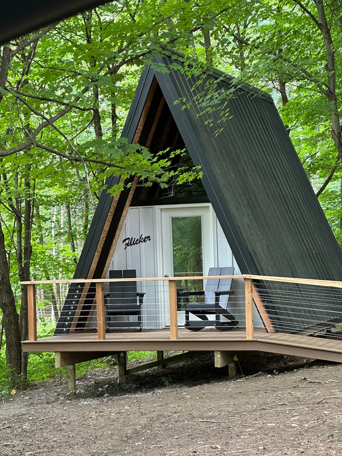 An A-frame cabin is nestled among lush trees, featuring a dark exterior and a welcoming porch with two black chairs. A wooden railing surrounds the deck, providing a cozy outdoor space to enjoy the serene wooded surroundings.