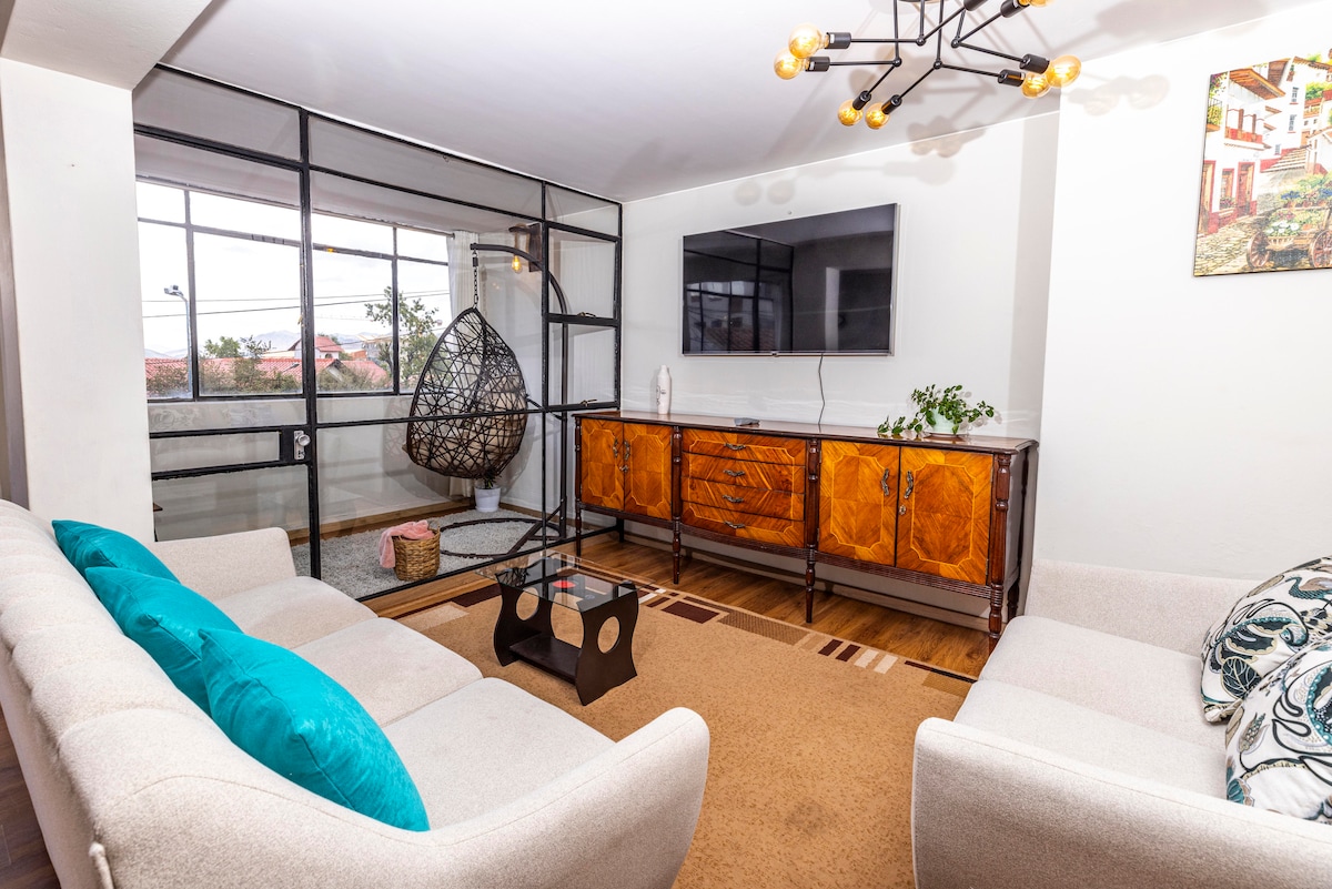 A bright living area features a light-colored seating arrangement with a soft rug underfoot. A modern television is mounted on the wall opposite a decorative cabinet. A hanging chair adds a unique touch, while large windows provide ample natural light and views of the surroundings.