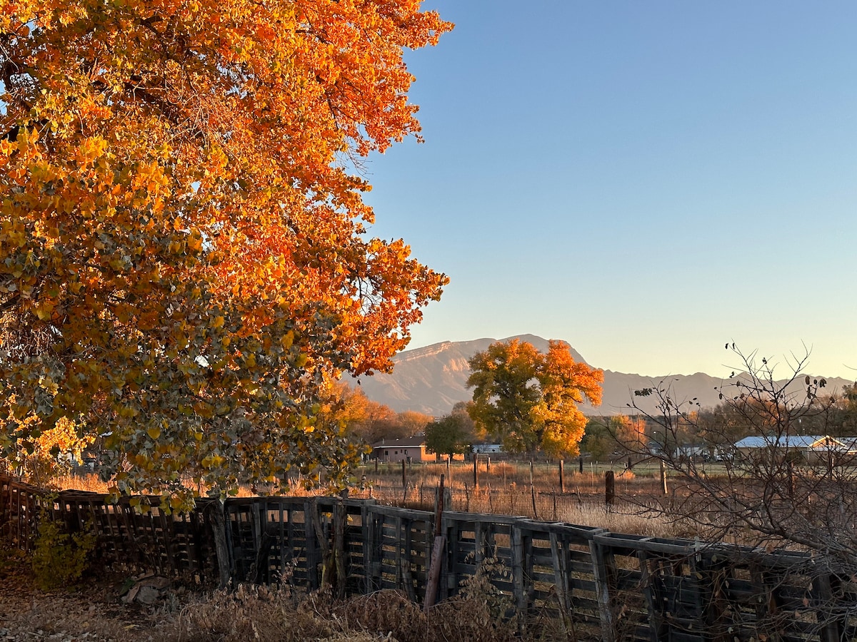 A vibrant autumn scene features trees adorned with orange foliage against the backdrop of the Sandia Mountains. The foreground showcases a rustic wooden fence, with expansive fields gently fading into the distance, creating a peaceful rural atmosphere.