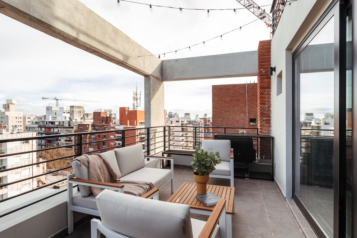 An expansive outdoor terrace is furnished with light-colored seating and a wooden coffee table, offering views of the cityscape. Soft lighting from overhead string lights creates a relaxed ambiance, while a potted plant adds a touch of greenery to the space.