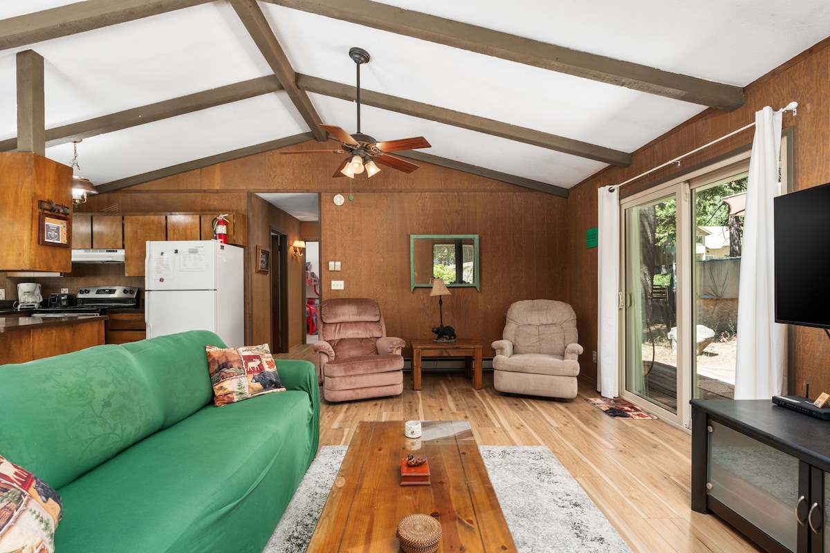 An open living area is highlighted by warm wood paneling and a sloped ceiling. A green sofa is placed in front of a wooden coffee table, accompanied by two light-colored recliners. Natural light filters in through sliding glass doors, leading to the outdoor space.