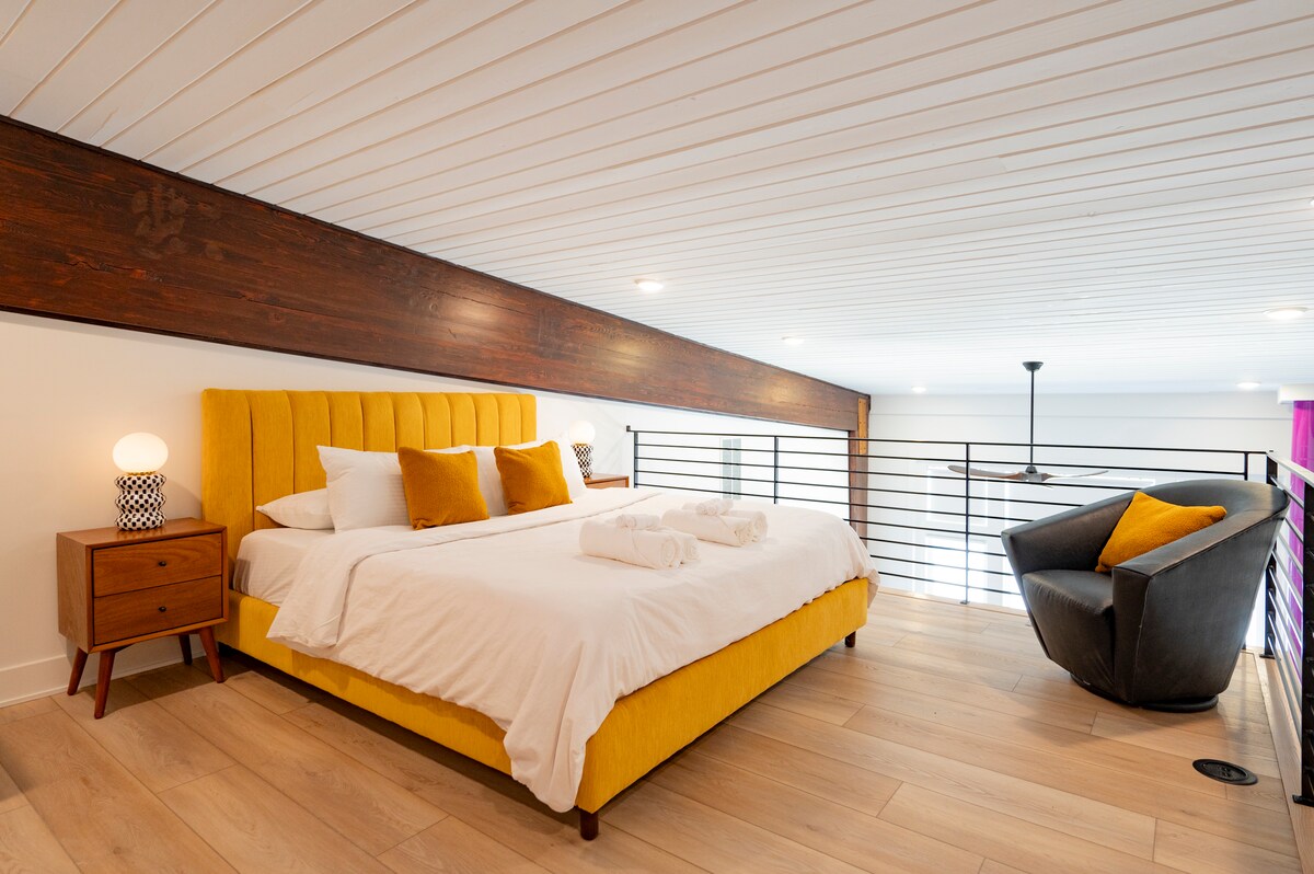 A lofted bedroom features a California King bed with crisp white linens and decorative pillows. Soft lighting is provided by bedside lamps on stylish nightstands. A cozy seating area with a black accent chair complements the warm yellow tones of the bed frame and cushions.