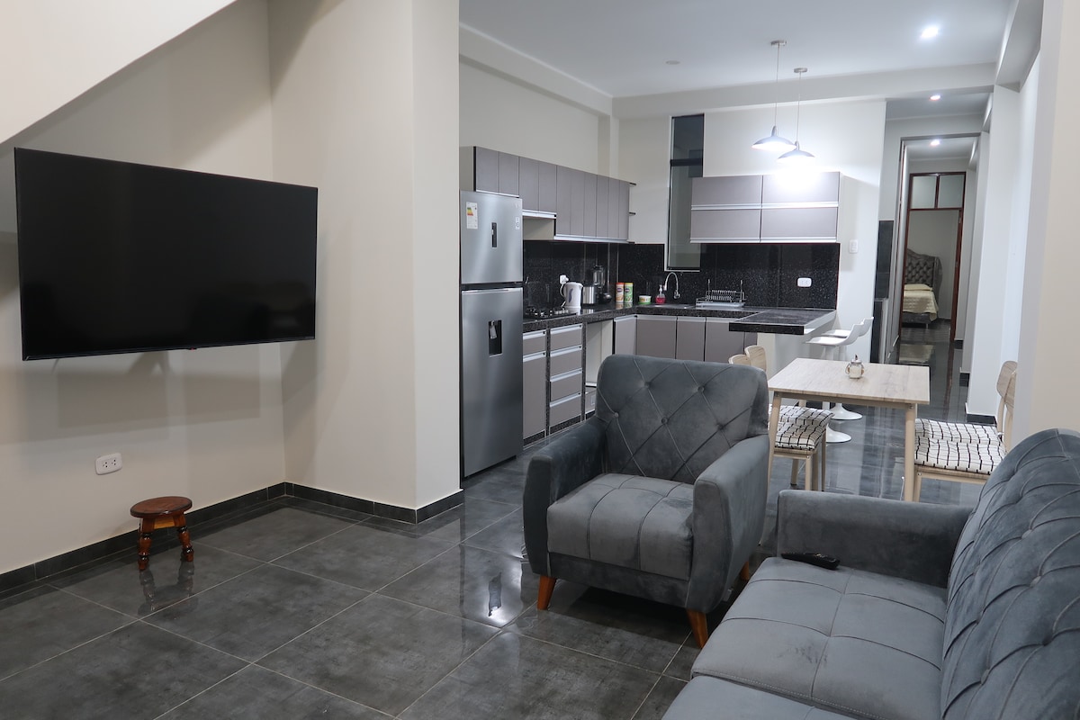 An open living area is presented with a modern gray sofa and a small dining table, positioned near a sleek kitchen. A flat-screen TV is mounted on the wall, while kitchen appliances are neatly arranged against dark cabinetry. The floor features glossy tile, enhancing the spacious feel.