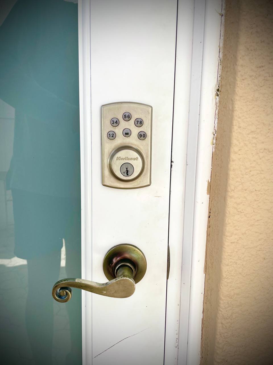 The image shows a close-up of a door handle and lock on a white door. The handle features an elegant design with a bronze finish. A rectangular nameplate is positioned above the handle, while the surrounding wall displays a light beige hue.