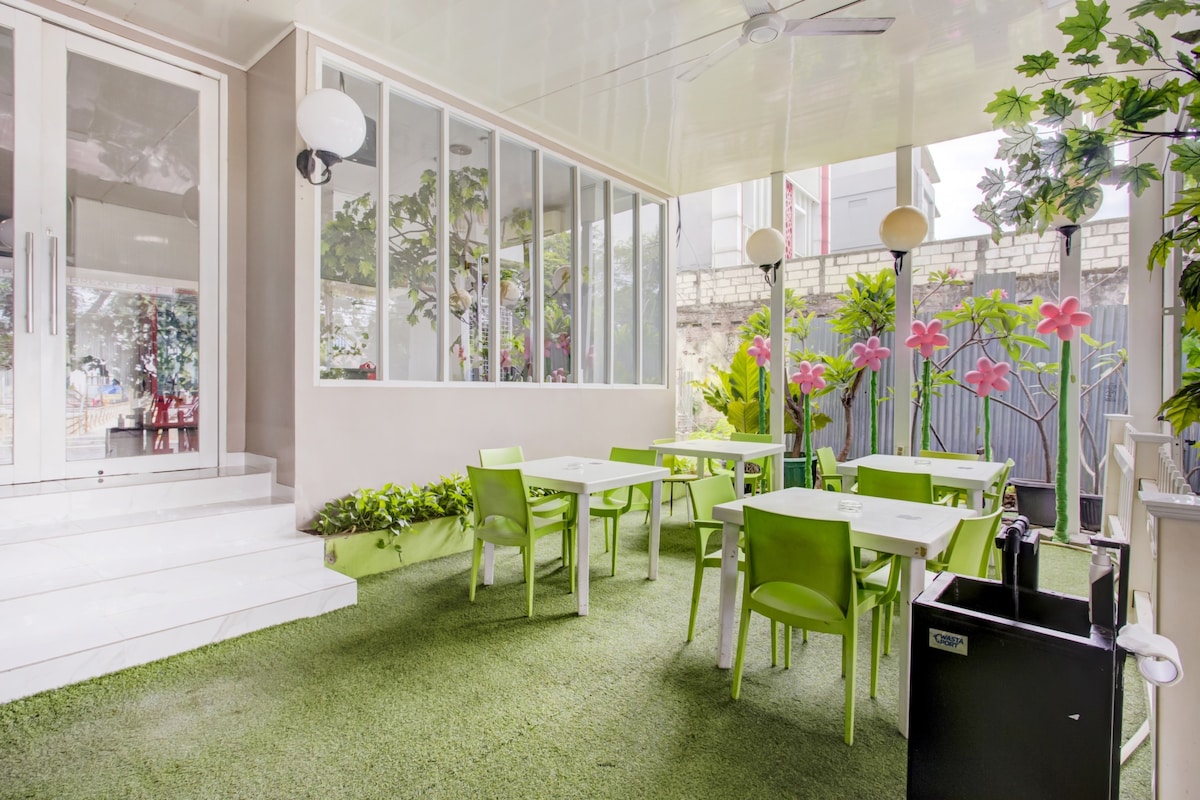An outdoor seating area is presented, featuring green tables and chairs positioned on artificial grass. Large glass walls allow natural light to enhance the space. Decorative plants and vibrant flowers contribute to a refreshing ambiance, creating a serene environment for relaxation.