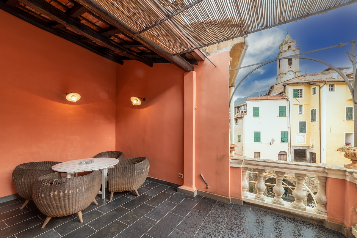 A shaded terrace area features comfortable round wicker chairs and a small table set against a backdrop of historic buildings with colorful façades. The space is framed by a trellis structure, providing a cozy spot for relaxation.