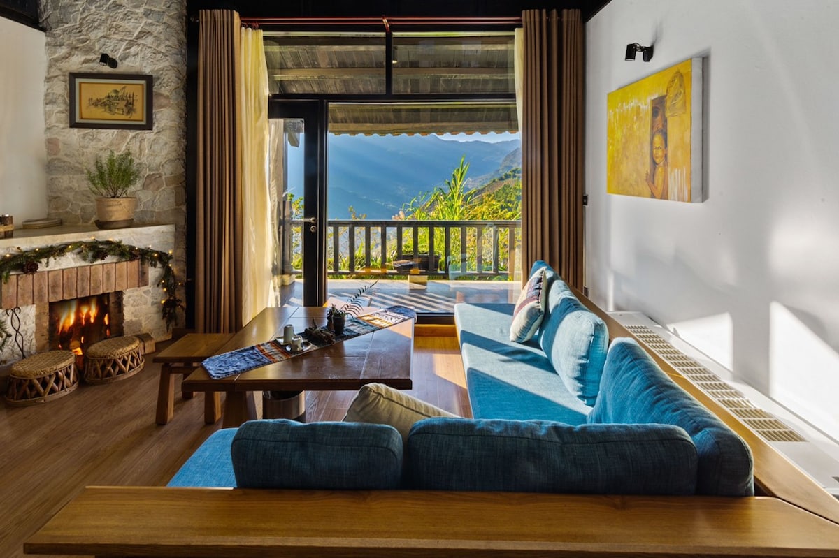 A spacious living area features a large blue sectional sofa and a wooden coffee table. Large glass doors allow natural light in, offering views of the surrounding mountains. A stone fireplace adds warmth to the space, while decorative elements enhance the cultural ambiance.