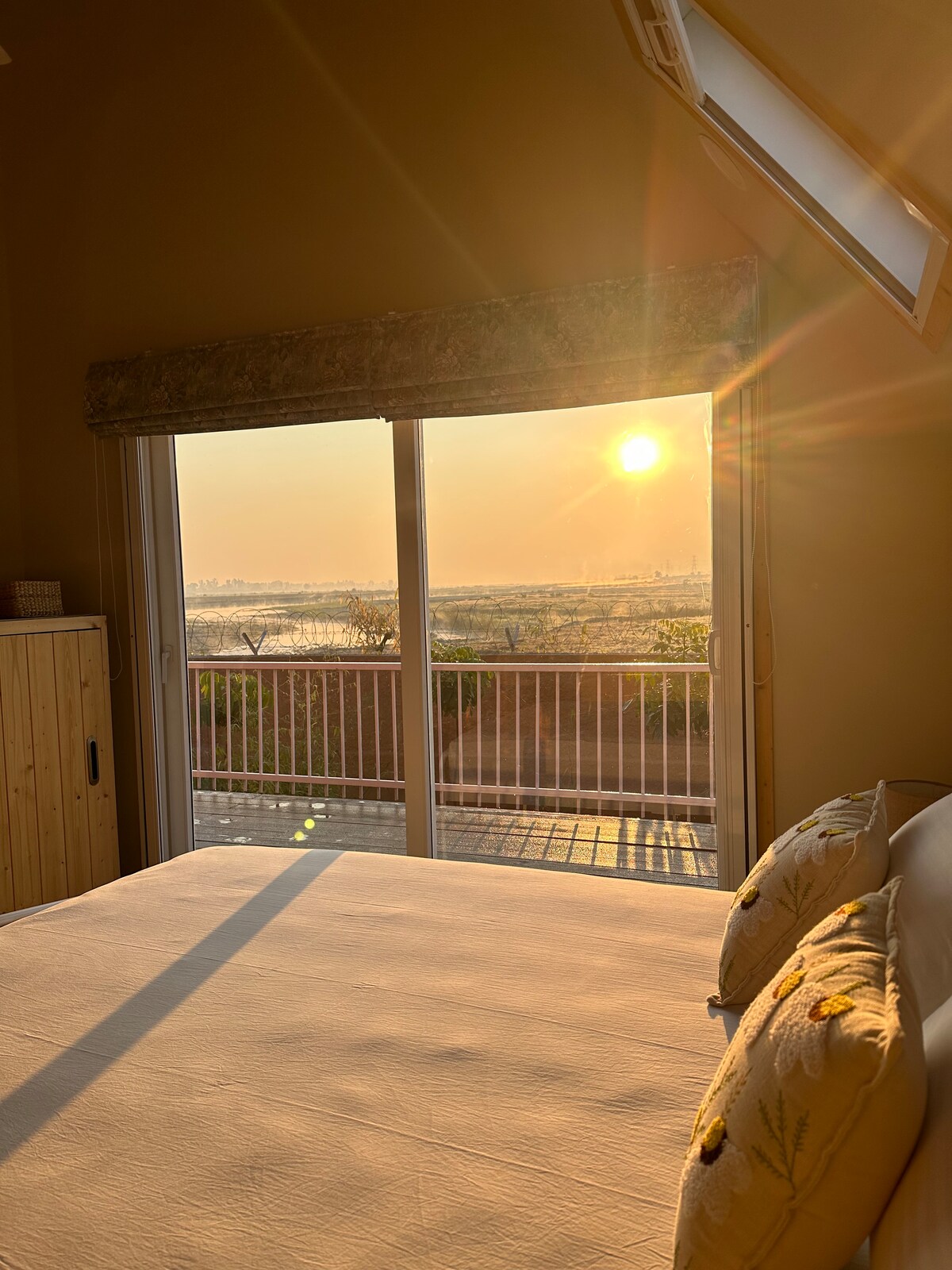 A serene bedroom is illuminated by warm sunlight streaming through large glass doors. A spacious bed with soft linens is positioned near the door, while a view of the river and surrounding nature is framed by the balcony railing.