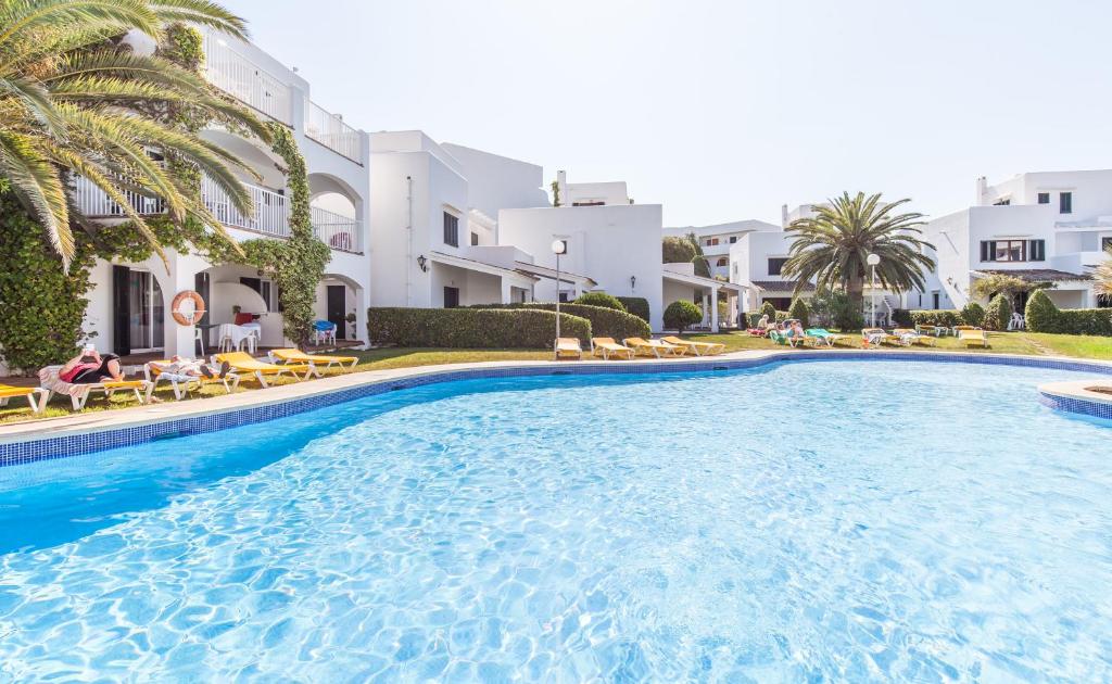 A clear blue swimming pool is flanked by lush green lawns and palm trees. Surrounding buildings feature a whitewashed design, with lounge chairs arranged along the poolside. Guests are seen enjoying the area, creating a relaxed atmosphere in the sunlit setting.