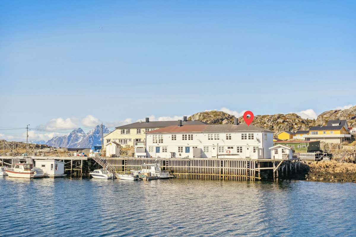 A view captures the exterior of StasBua, a waterfront building nestled against rocky terrain. The structure reflects a blend of historical and modern architecture, with boats docked nearby. Mountains are visible in the background, showcasing a natural setting that complements the tranquil atmosphere.