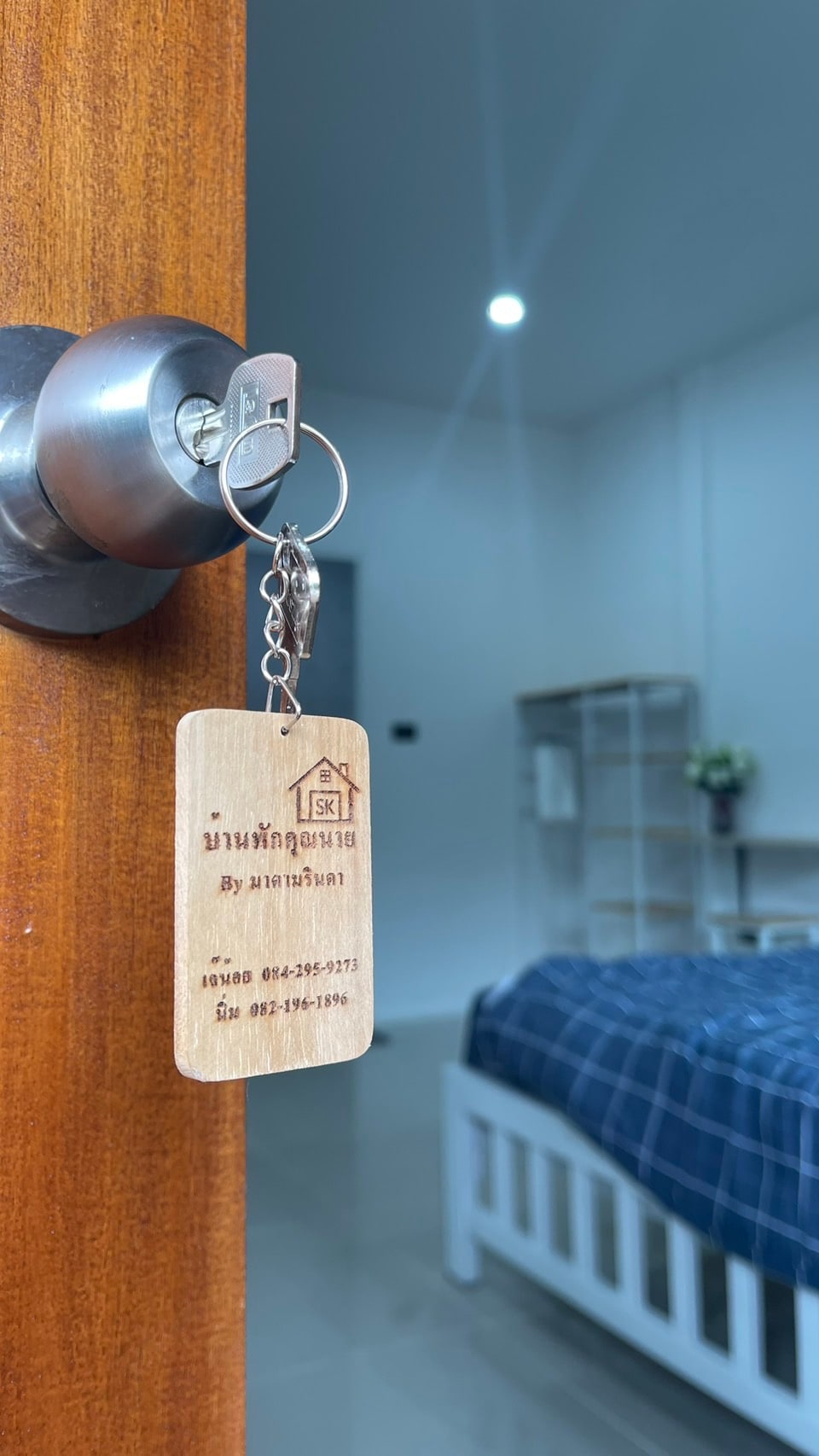 A wooden key tag hangs from a door knob, with a key inserted. The background reveals a tidy sleeping area featuring a white bed with a navy blue checkered blanket. Soft lighting illuminates the room, and a decorative arrangement of flowers can be seen on a shelf.