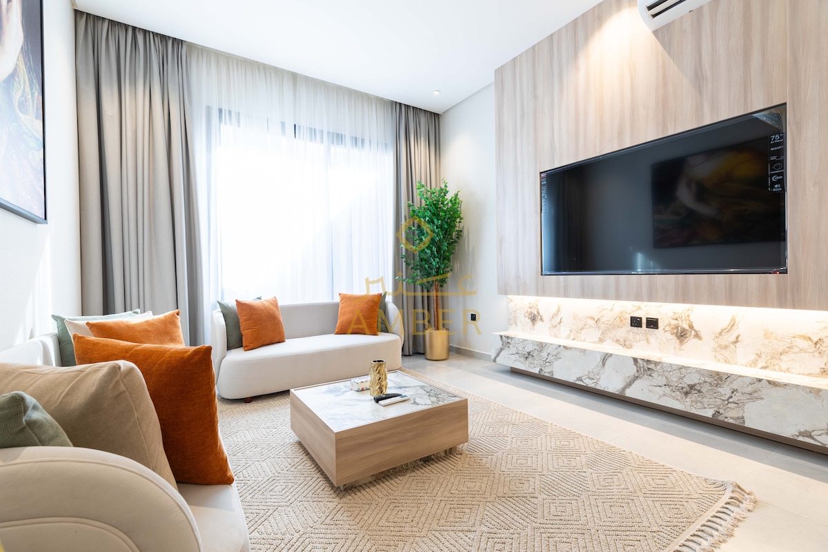 A modern living area is presented with a large flat-screen TV mounted on a wall, surrounded by a stylish media console. A soft rug lies beneath a low coffee table, complemented by a mix of cushions on the couch and accent chairs. Natural light filters through layered curtains.