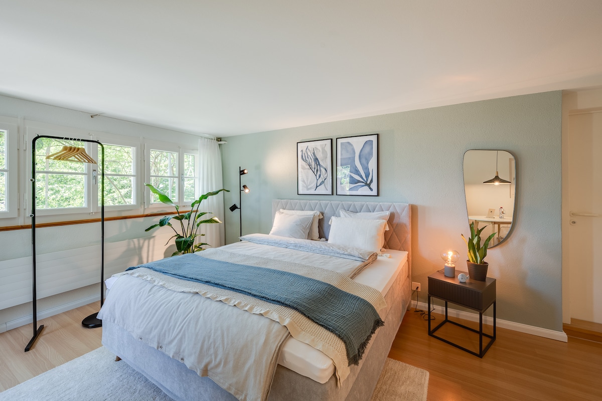 A spacious sleeping area features a comfortable bed with neutral-toned bedding and decorative pillows. Natural light floods through multiple windows, highlighting indoor plants. A minimalist mirror and a side table with a candle add subtle elegance to the modern decor.