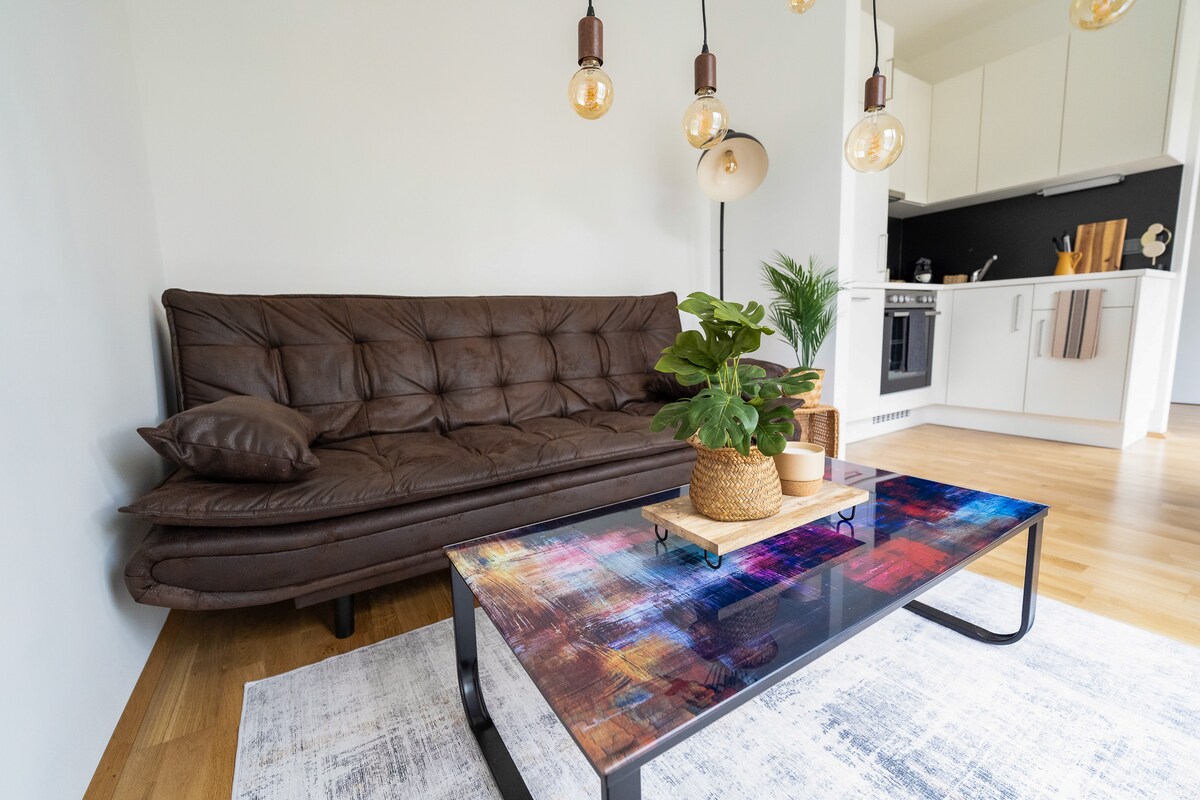 A cozy living area is highlighted by a brown leather sofa and a colorful glass coffee table. A small potted plant and decorative tray sit atop the table. Warm pendant lights hang from the ceiling, illuminating the space, which leads to a modern kitchen in the background.