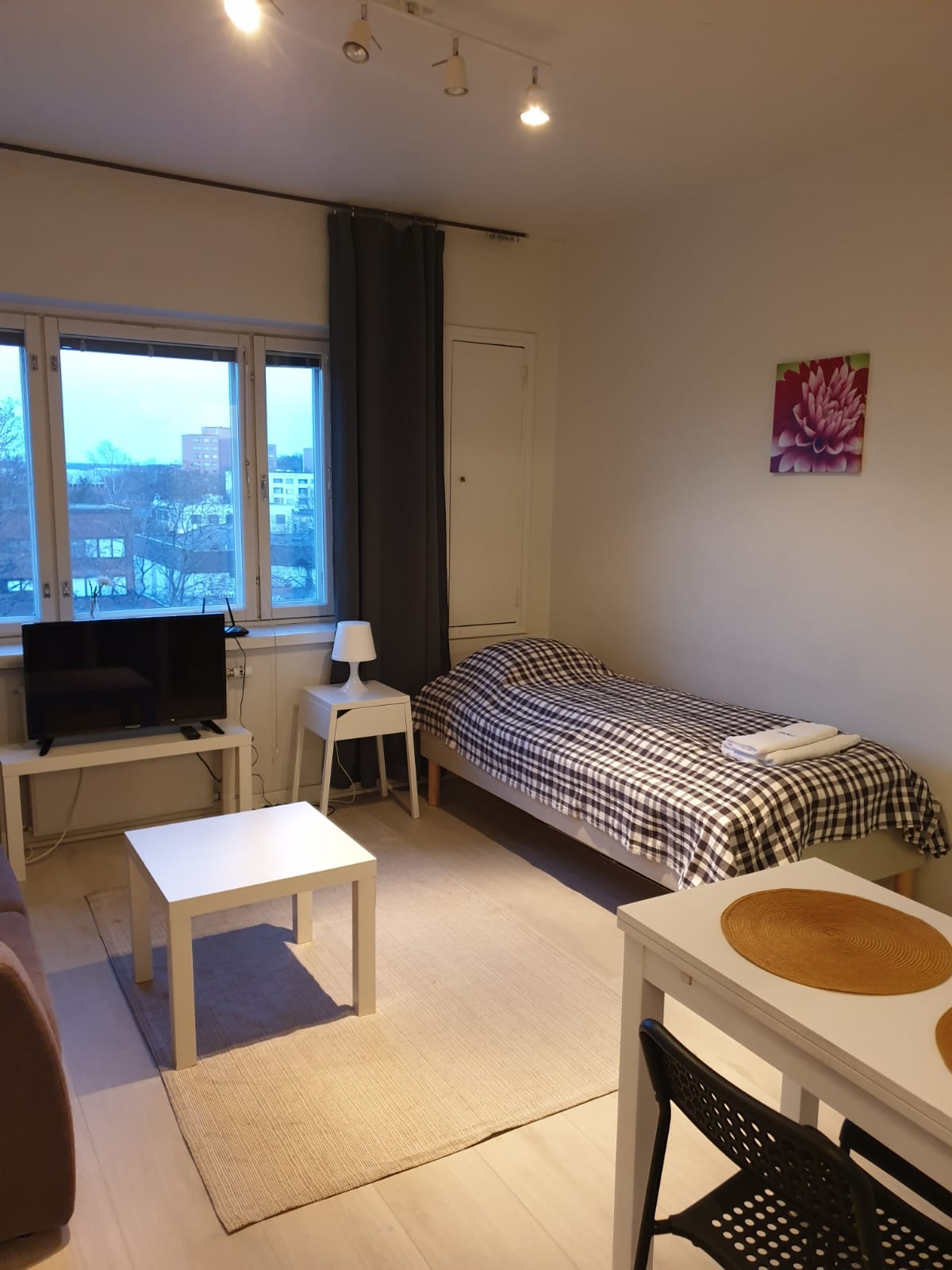An inviting studio space features a single bed dressed in a plaid coverlet and is positioned near a window. A small television stand and a light source complement the room. A dining table and chairs are located on the opposite side, with a textured area rug offering additional comfort.