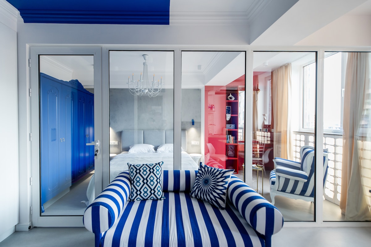 A stylish living area features a striped blue and white sofa, positioned in front of large glass doors that provide a view of the bedroom. The bedroom showcases a cozy bed against a grey accent wall, complemented by colorful furnishings and natural light streaming through the expansive windows.