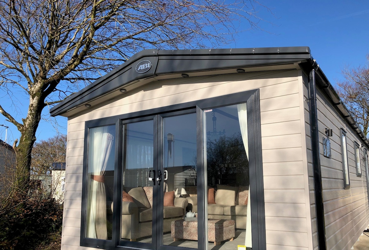 The exterior of the lodge features large glass windows, allowing natural light to fill the interior. The entrance is adorned with welcoming curtains, and a seating area with comfortable furnishings is visible inside. Surrounding trees contribute to the serene atmosphere.