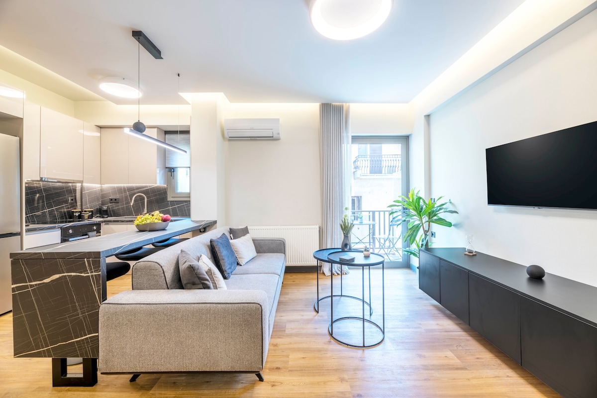 A modern living area features a light grey sofa adorned with plush cushions, positioned next to a round black coffee table. A wall-mounted TV and sleek cabinetry complement the minimalist decor. Natural light filters through large windows leading to a small balcony, enhancing the open atmosphere.