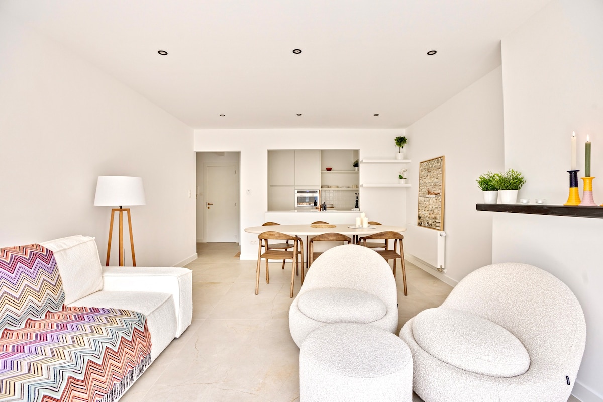 A bright living area features a soft sectional sofa with patterned upholstery and two rounded armchairs. A wooden dining table is set for four, with sleek, minimalistic decor enhancing the modern kitchen backdrop. The space is airy, illuminated by recessed lighting and natural light.