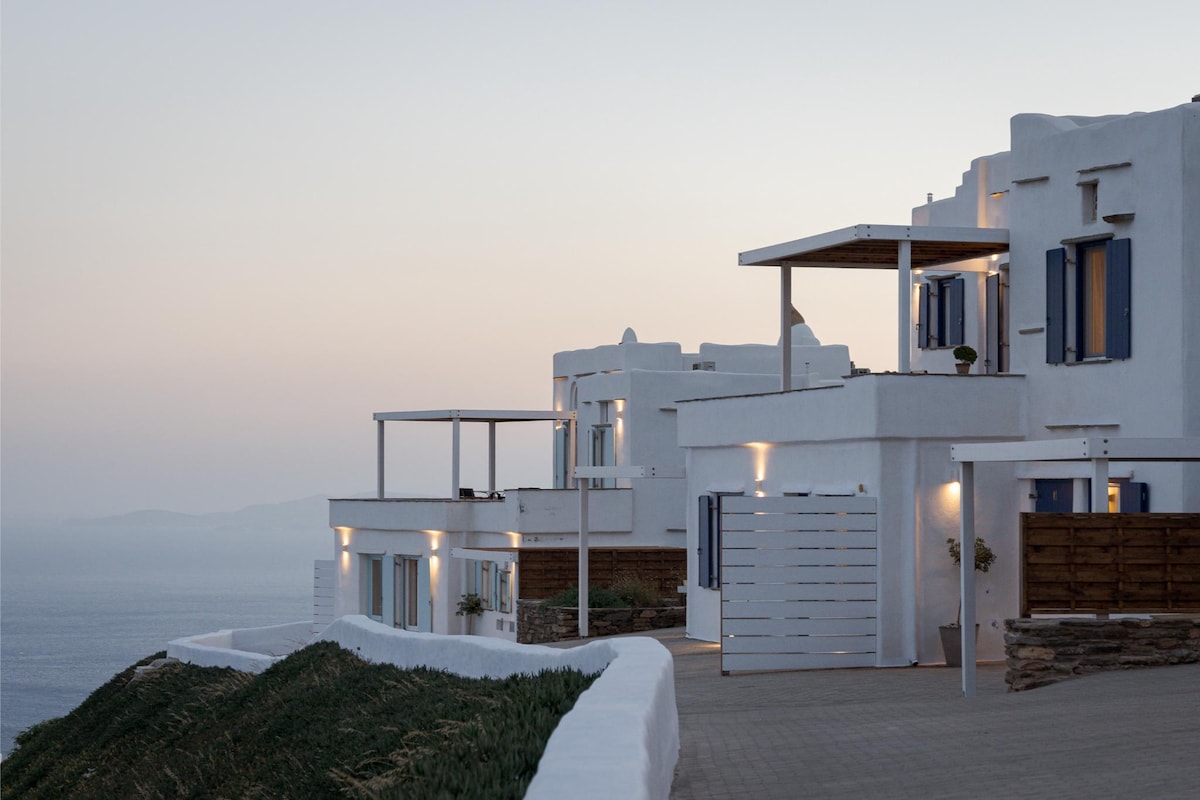 A collection of Cycladic-style apartments is displayed against a tranquil sunset sky. The white structures with blue shutters are positioned on a coastal hillside. The outdoor areas are illuminated softly, enhancing the serene atmosphere. The sea is visible in the background, completing the scenic view.