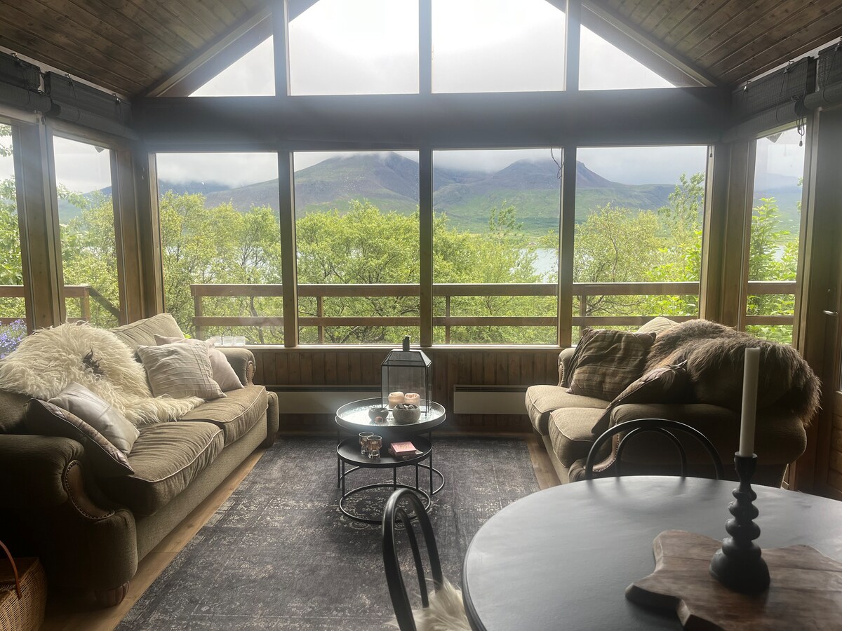 A spacious sunroom features large windows offering expansive views of surrounding mountains and greenery. Comfortable sofas are arranged around a circular coffee table, with a light rug anchoring the seating area. A small dining table is visible in the corner, enhancing the inviting ambiance.