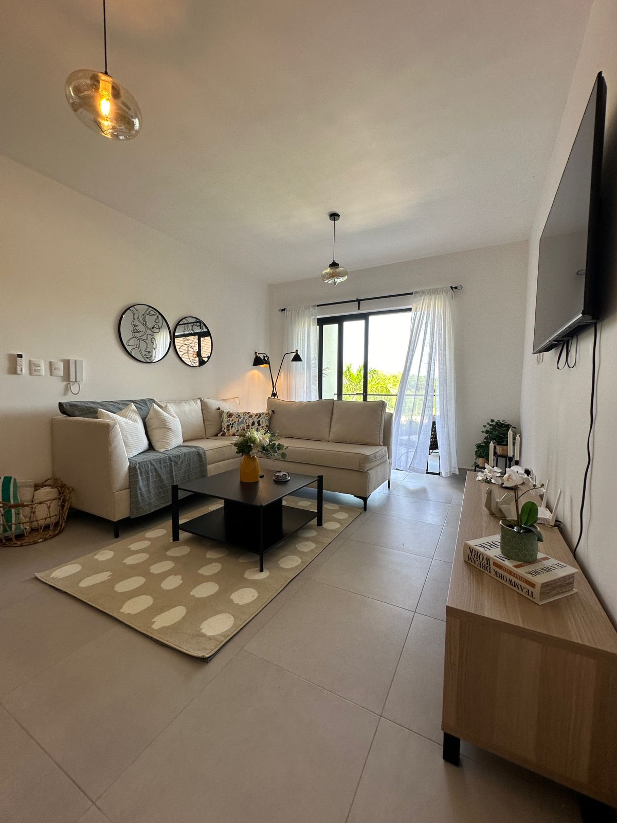 The living area features a comfortable sofa paired with decorative pillows, all arranged around a low coffee table on a chic patterned rug. Natural light floods in from the balcony door, complemented by modern lighting fixtures and tasteful wall decor.