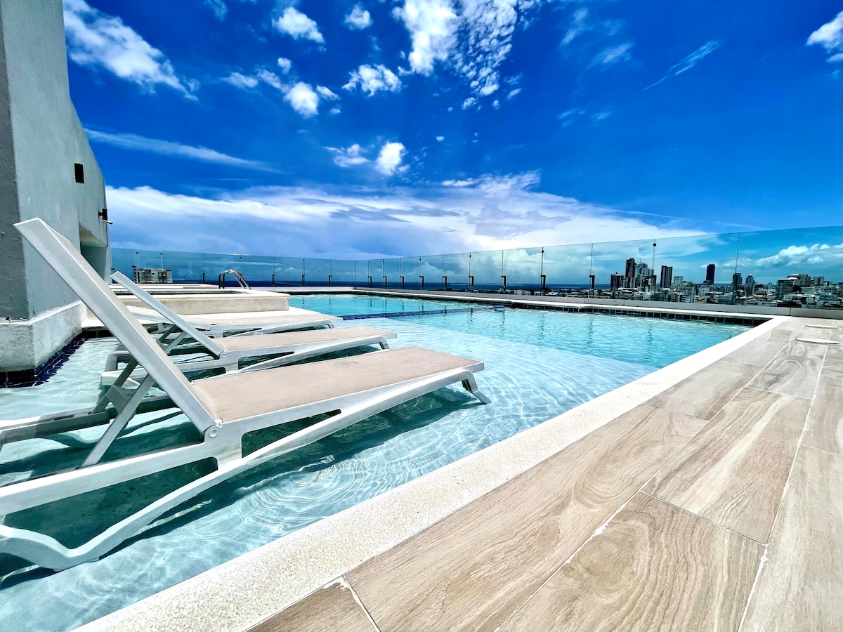 A rooftop pool area is depicted under a clear blue sky, featuring lounge chairs placed alongside the water. The pool's surface reflects the clouds, while the city skyline is visible in the background, adding a sense of urban tranquility.
