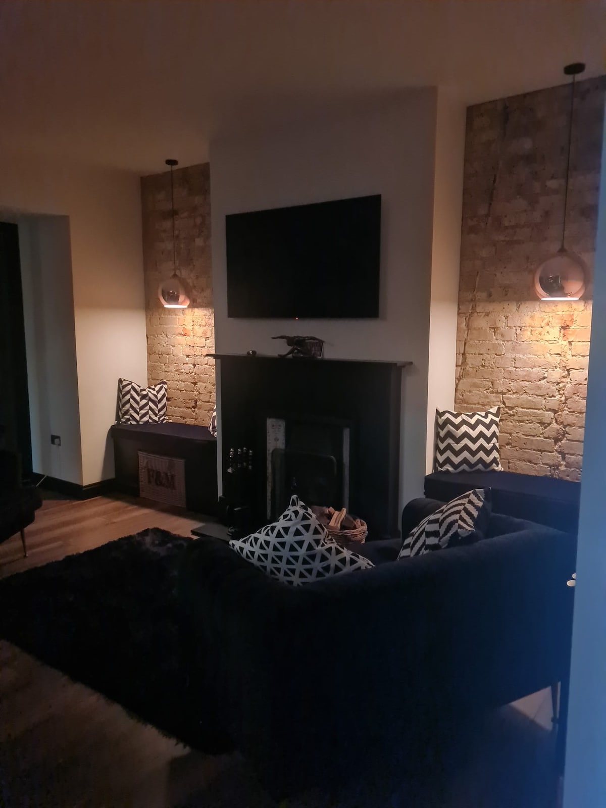 The snug lounge features exposed brick walls with two pendant lights illuminating a cozy seating area. A dark sofa with patterned cushions is positioned in front of a traditional fireplace. A flat-screen television is mounted above the fireplace, providing a modern touch.