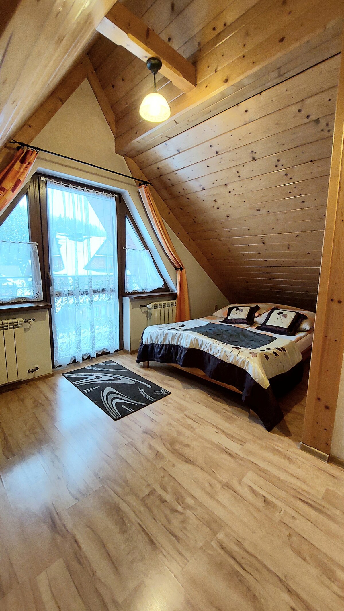 A cozy room features wooden walls and sloped ceilings, providing a warm atmosphere. A spacious bed is positioned against a window draped with sheer curtains. Natural light floods the space, illuminating the wooden floor and a small area rug.