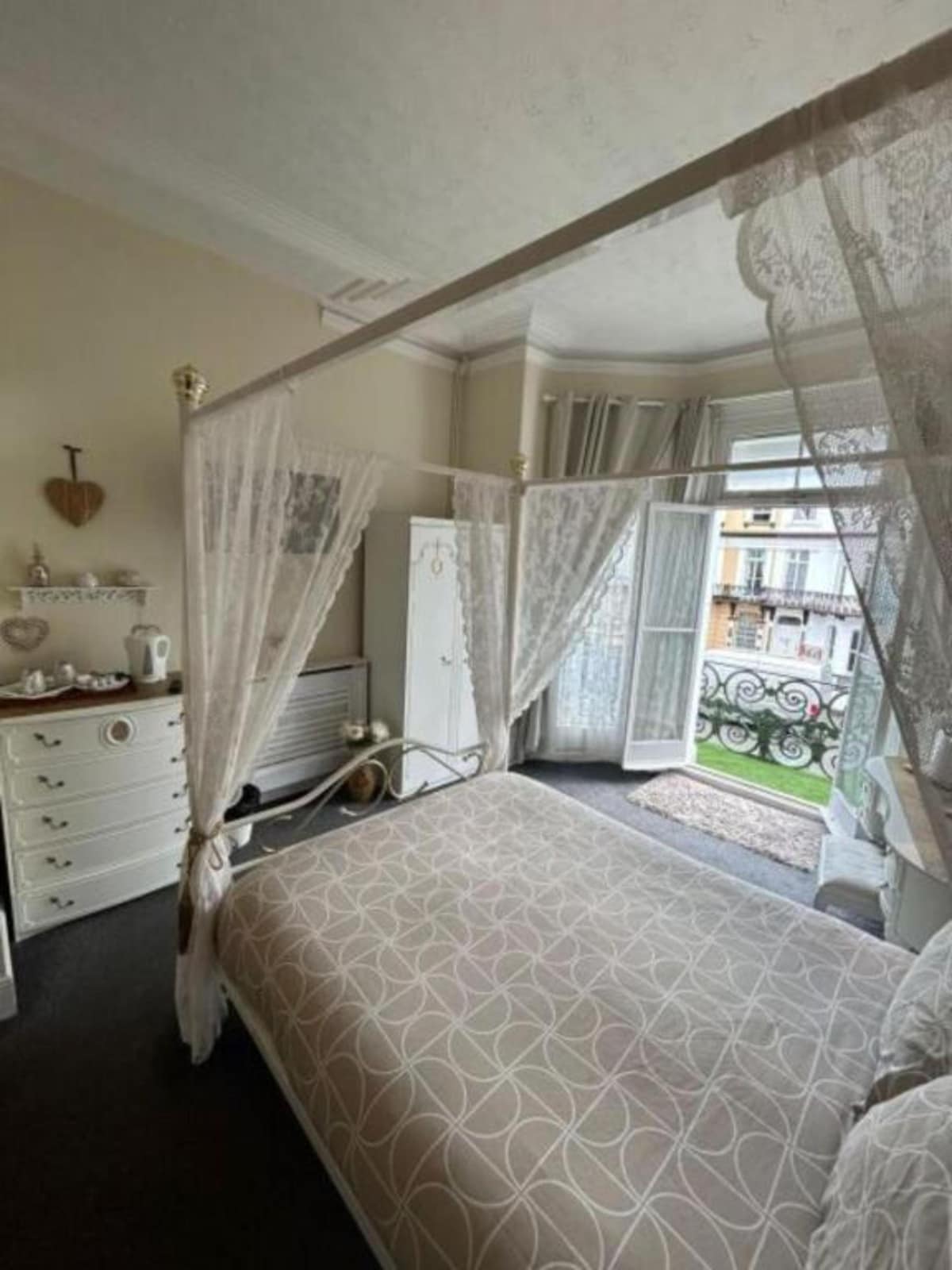 A serene bedroom features a canopy bed adorned with sheer drapes. A light-colored comforter lies across the bed, while large windows allow natural light to illuminate the space. A small balcony with decorative railing can be seen in the background, offering an outdoor view.
