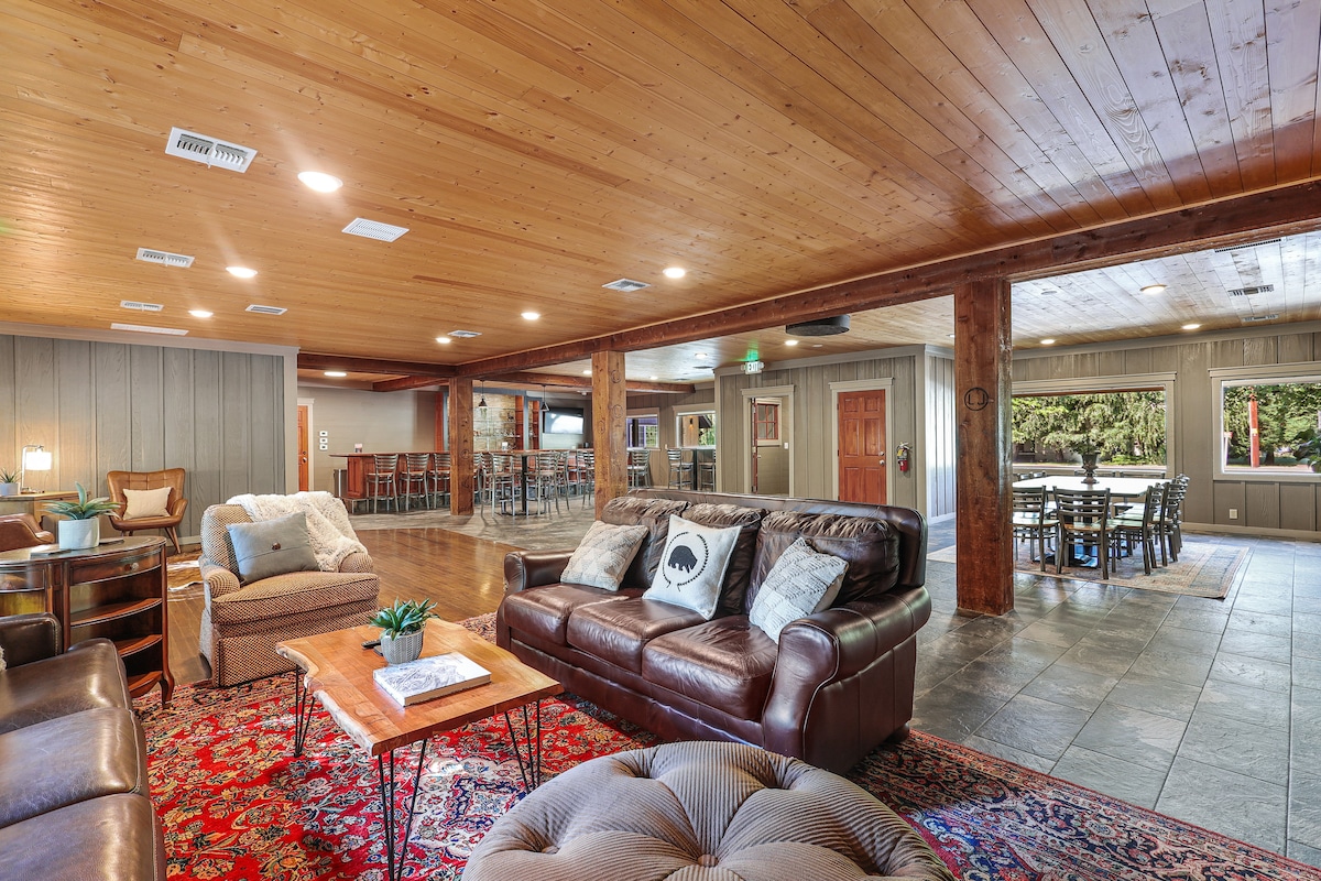 An open communal area features natural wood ceilings and a rustic charm. Brown leather seating arrangements are complemented by a patterned area rug. Large windows allow natural light, while a dining area with wooden tables and chairs is visible in the background.