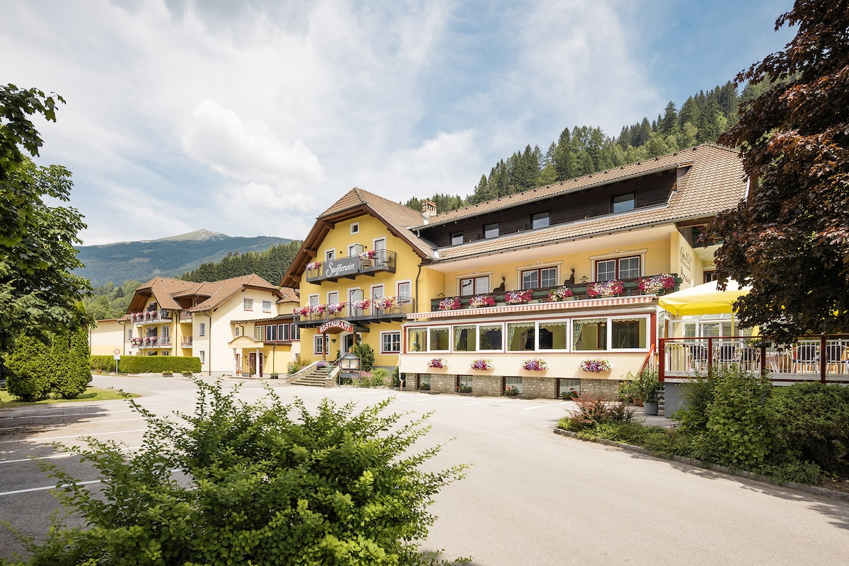 A charming multi-level building with a yellow facade is surrounded by green foliage and mountains. Flower boxes adorn the windows, enhancing the exterior. A spacious parking area is visible in front, while a covered terrace offers seating and shade.